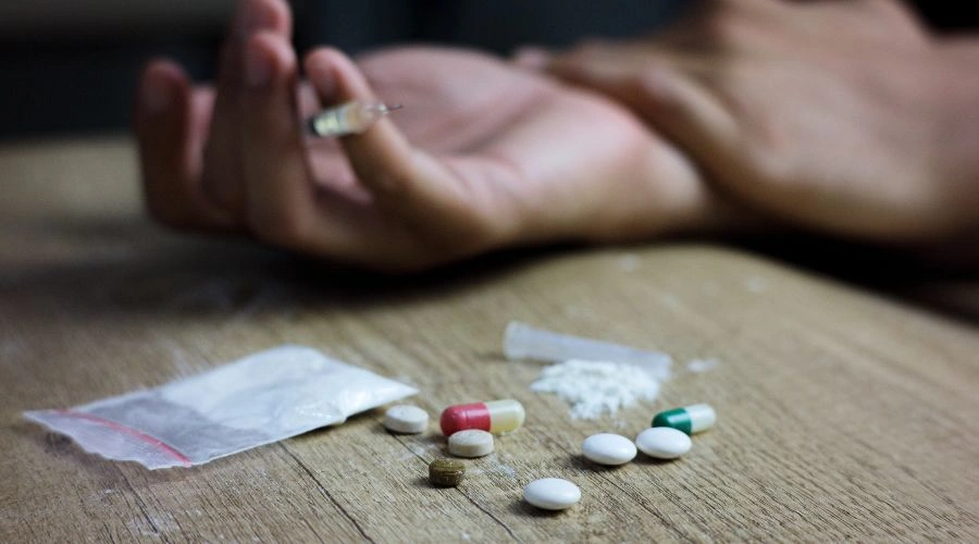 Hand holding a syringe and needle with pills on a table displaying dPost-Traumatic Stress Disorder Impact Substance Abuse