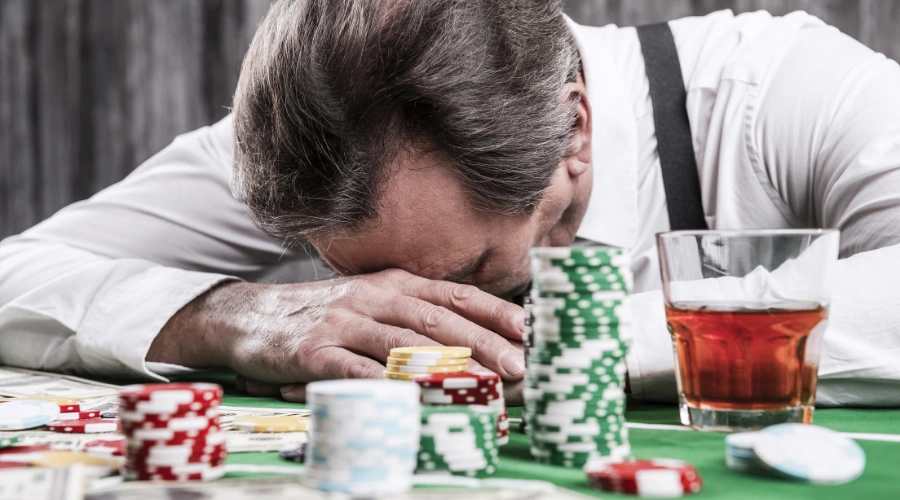 Male sitting with his face on a table in anguish, sitting in front of a glass of liquor and poker chips