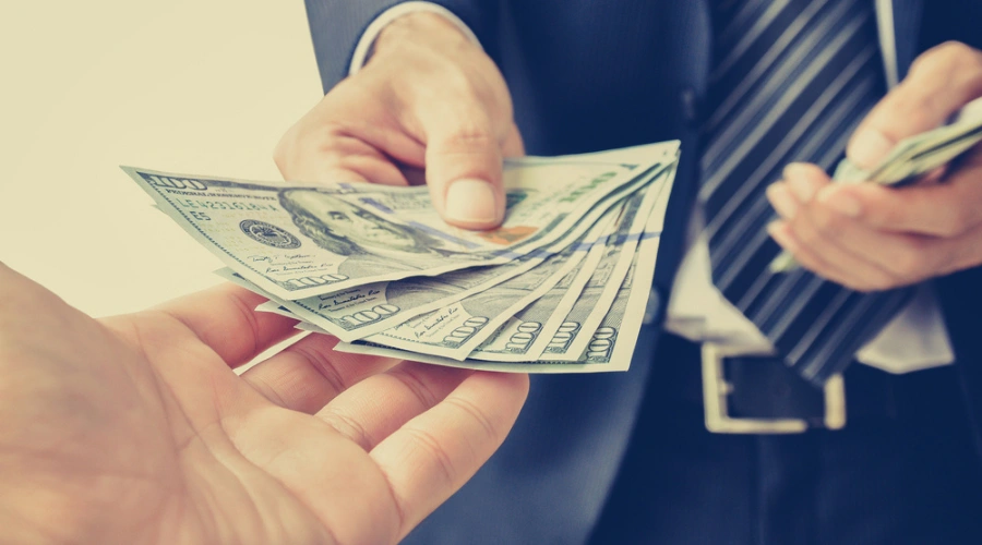 Image of a man in a suit giving several $100 bills to the hand of another person - Difference Between Enabling and Supporting an Addicted Sibling