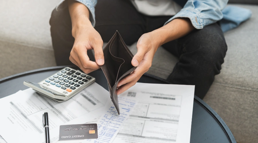 Image of a man holding an open, empty wallet, unable to pay a stack of mounting unpaid bills