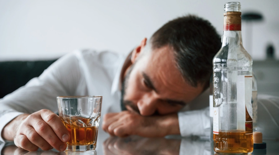 Image of man sitting with his head on a table beside an almost empty bottle of a liquor and a half empty glass of liquor in his hand - Gambling withdrawal and mental health