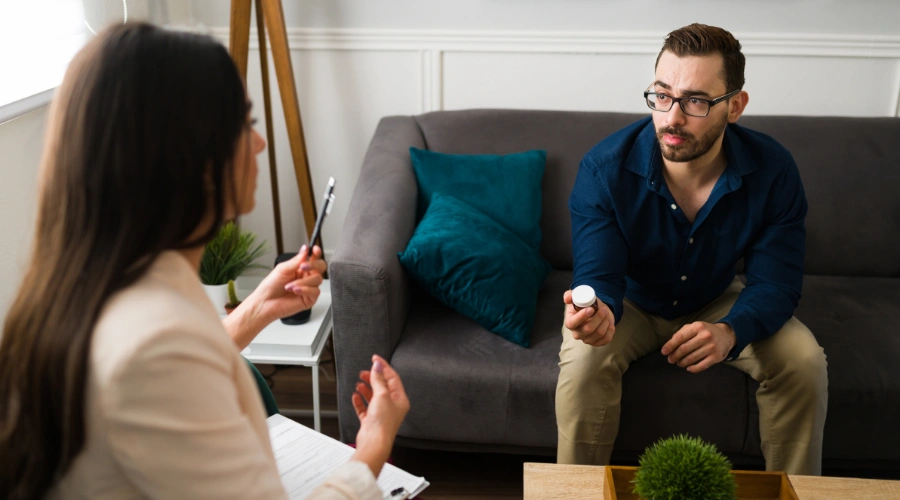 Image of male patient sitting on a sofa and holding a bottle of medication as a counselor beside him looks on