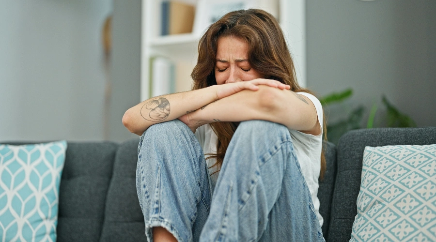 Image of a woman in deep anguish, crying and sitting on a sofa with her arms on her knees experiencing a trauma meltdown
