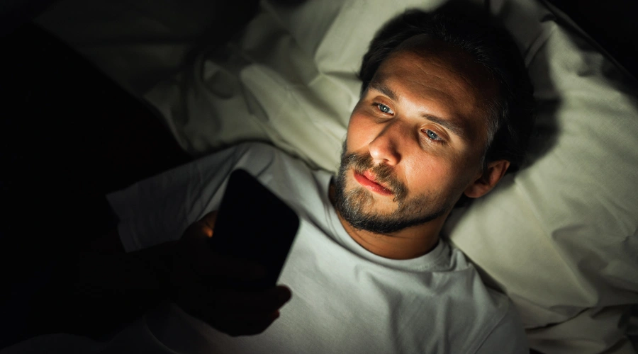 Image of a man lying in bed at night using a smartphone, with the screen’s glow illuminating on his face, showing how late-night social media use affects sleep quality