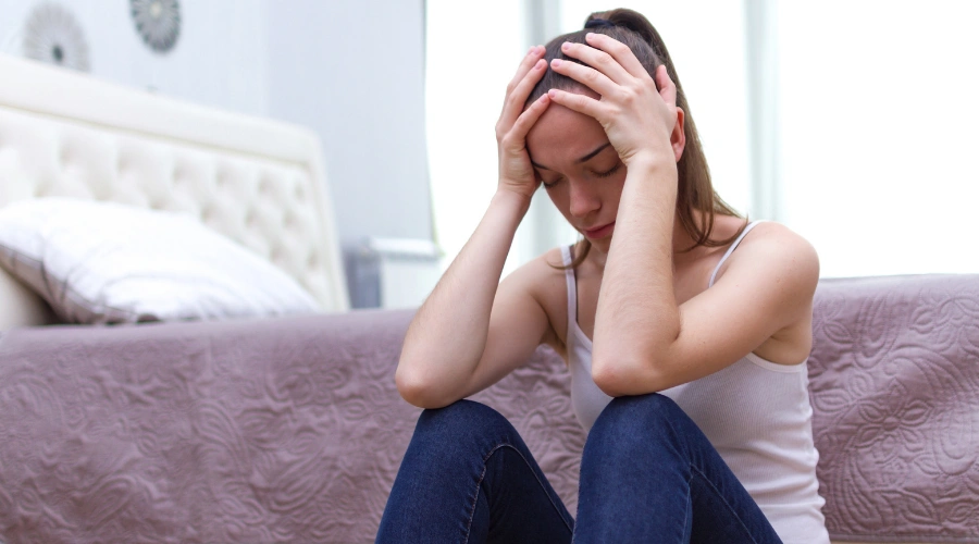 Image of a woman sitting on the floor beside her bed, holding their head in stress, showing how chronic stress affects the nervous system and emotional balance