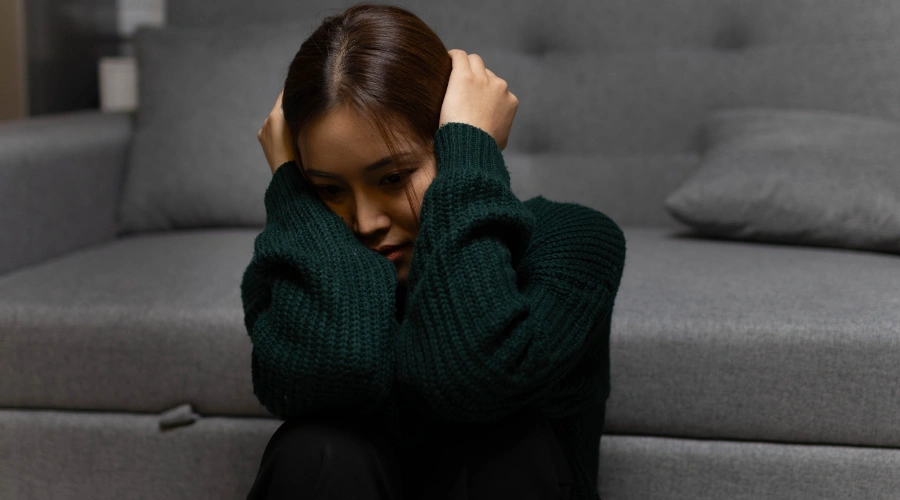 Image of female sitting alone on the floor in front of a sofa in deep thought, displaying the emotional and mental health effects of childhood trauma
