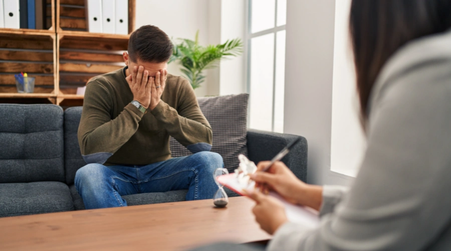 Image of a man suffering with anxiety management holding his hands over his face as a therapist looks on writing down information