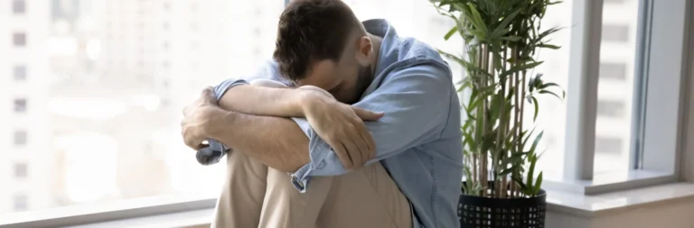 Image of a man sitting on the floor holding his face next to his elbows, against his knees - Why Does My Anxiety and Depression Come in Waves - Icarus Behavioral Health Nevada
